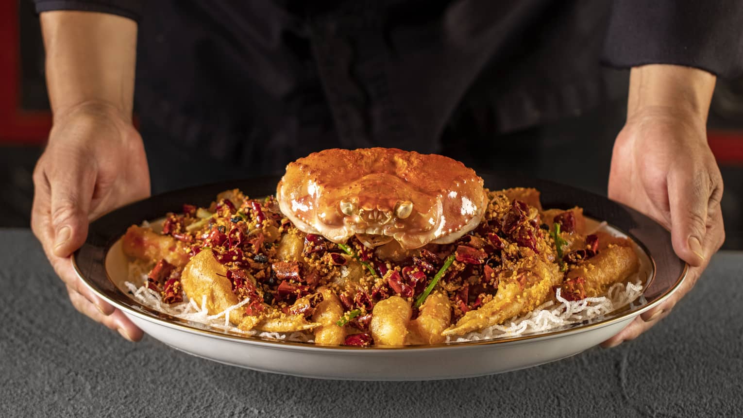 Two hands hold dish of whole crab on bed of crispy rice noodles