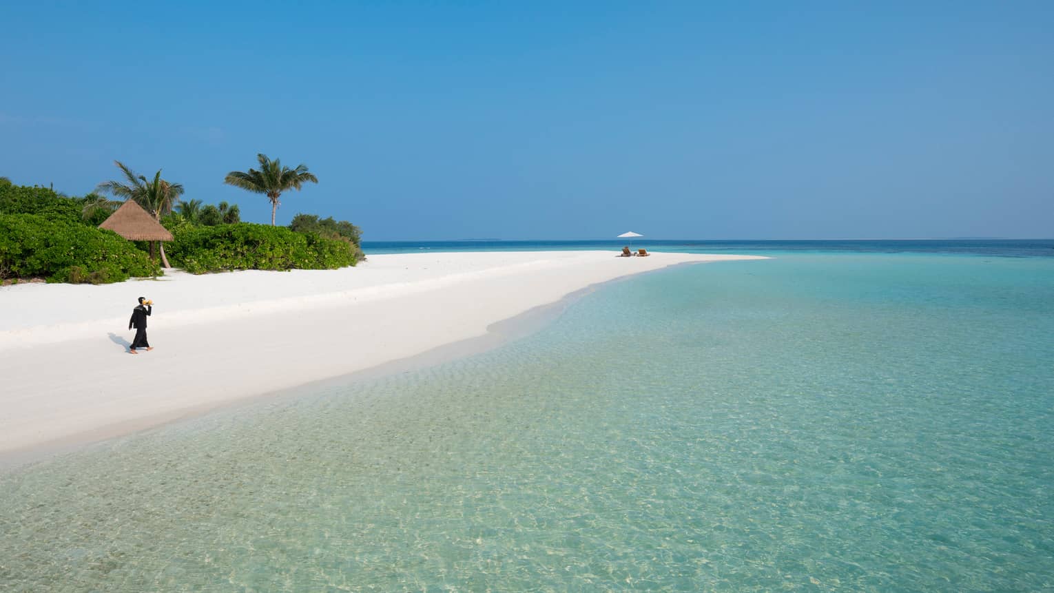 Hotel staff carries tray across long white sand beach to couple in lounge chairs by lagoon