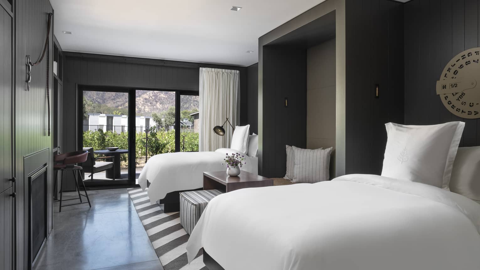 Luxury resort room with two beds, dark paneled walls, seating nook, decorative wall art, striped rug, fireplace and patio with vineyard and mountain view