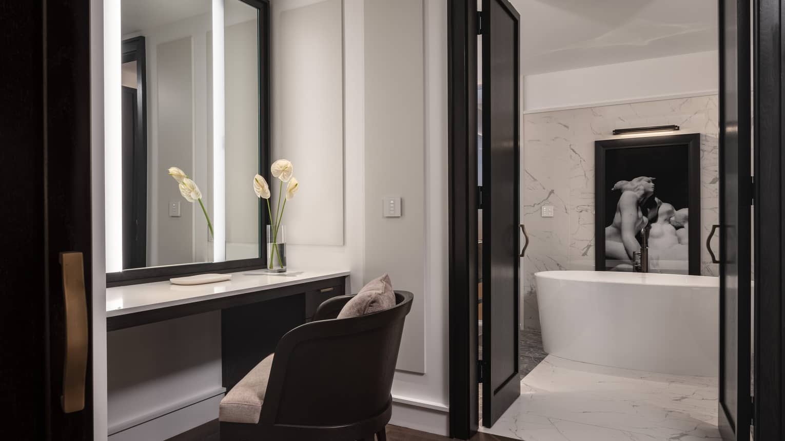 A modern bathroom houses a freestanding bathtub framed by marble walls. Adjacent is a vanity area with a chair, backlit mirror and a vase holding white flowers.
