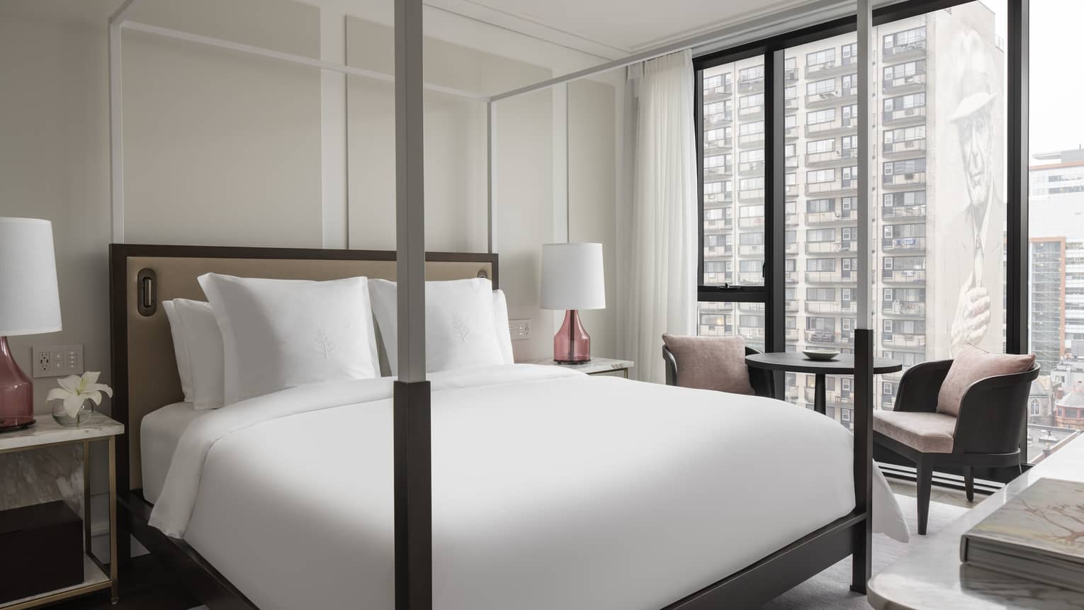 Four-post bed with white linens framed by marble nightstand topped with lamps on each side sits next to a small table with two chairs and floor-to-ceiling windows