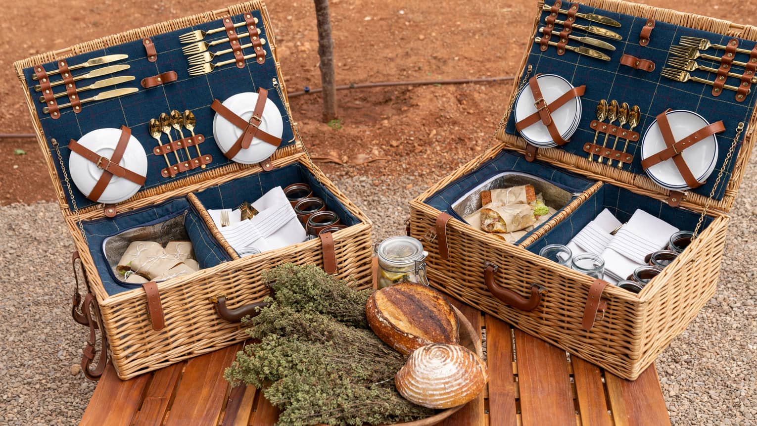Plates and gold cutlery secured to the lids of picnic baskets containing parchment-wrapped sandwiches, napkins, and glasses.