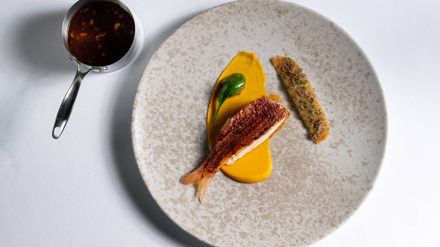 Red Mullet Bourguignon with Smoked Eel, Pearl Onion and Potato Galette on tan stone plate with measuring cup on side