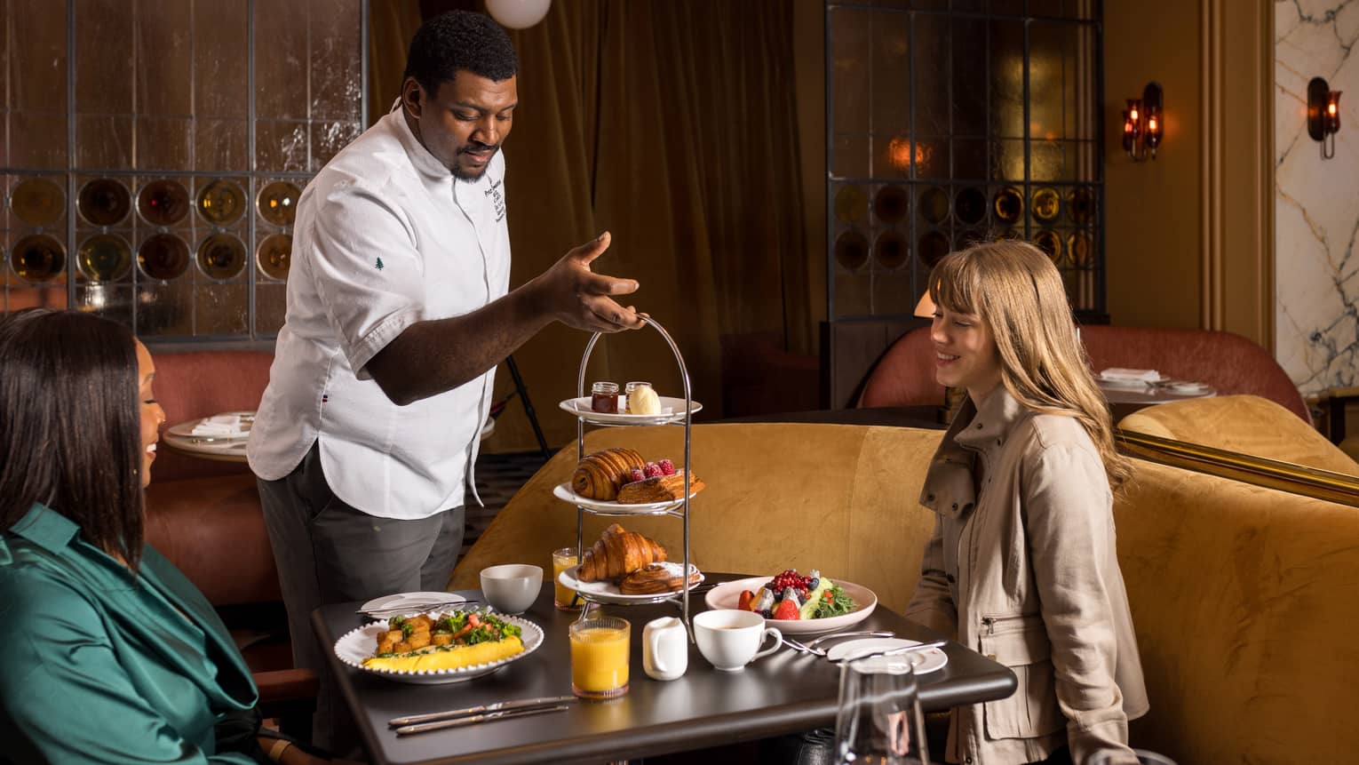 A person wearing a chef?s coat placing a three-tier display of sweets on a dining table