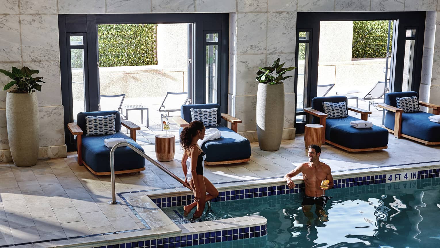 A woman steps down into an indoor pool towards a man lounging while holding a yellow cocktail