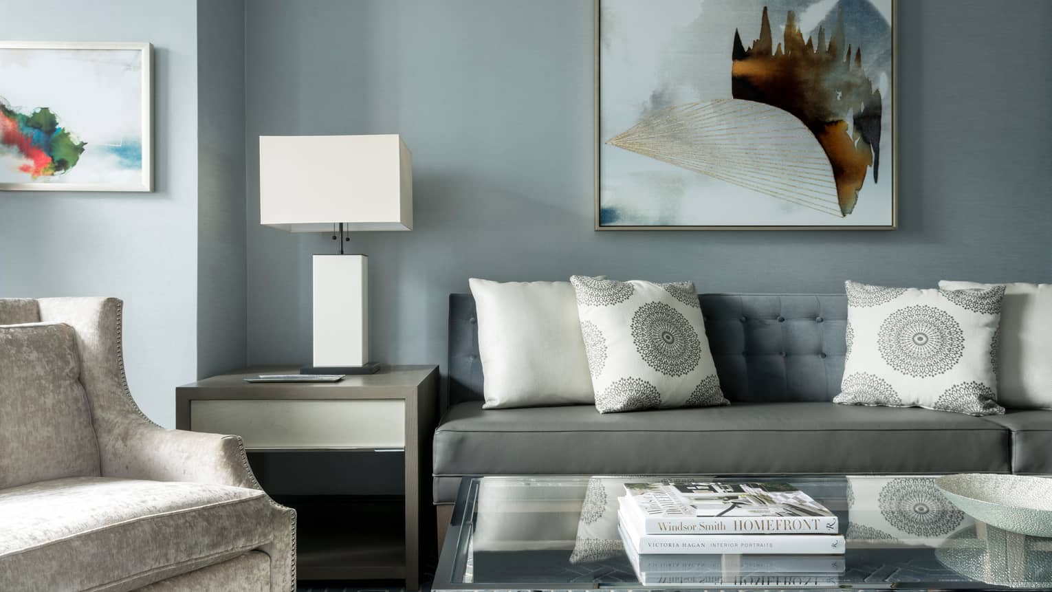 Midtown Suite long grey sofa with white accent pillows below abstract painting, table, lamp, armchair