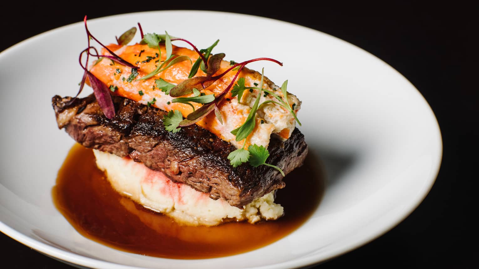 Close-up of grilled steak topped with lobster and fresh herbs, on bed of mashed potato with jus