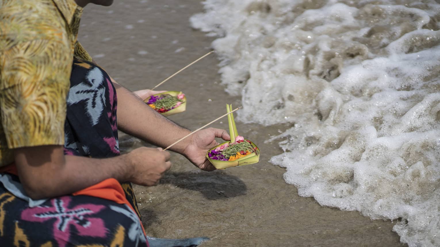 Kneeling at the edge of frothy water, a person in colourful ceremonial dress offers incense and a small woven box of flowers.