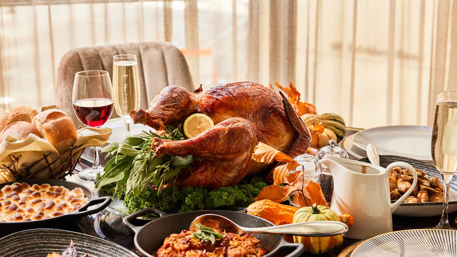 Table set with a roasted turkey, bread rolls, various side dishes, glasses of wine and champagne, and decorative gourds.