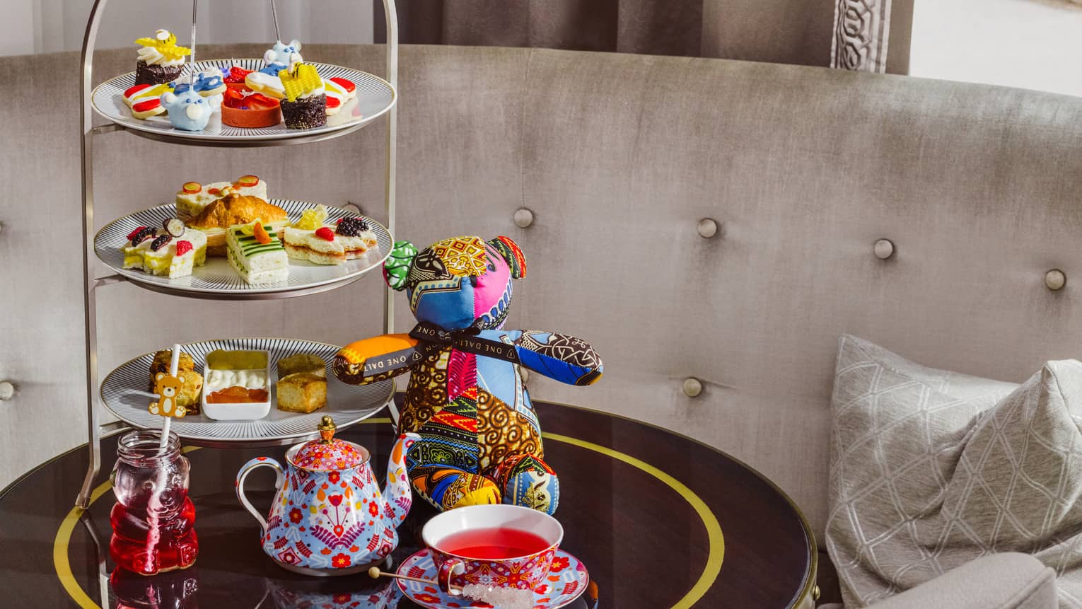 A small tabletop next to upholstered seating holds a decorative teapot and cup, a display of small bites, and a colourful stuffed teddy bear.
