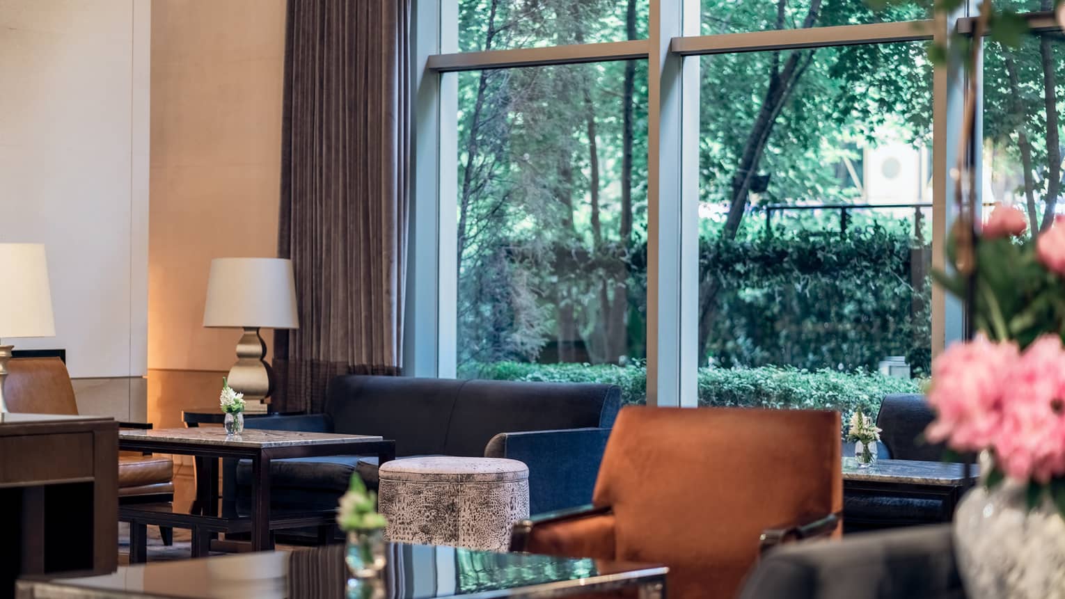 Glass-topped tables, blue couch, tan accent chair, floor-to-ceiling windows with open curtains displaying green foliage outside