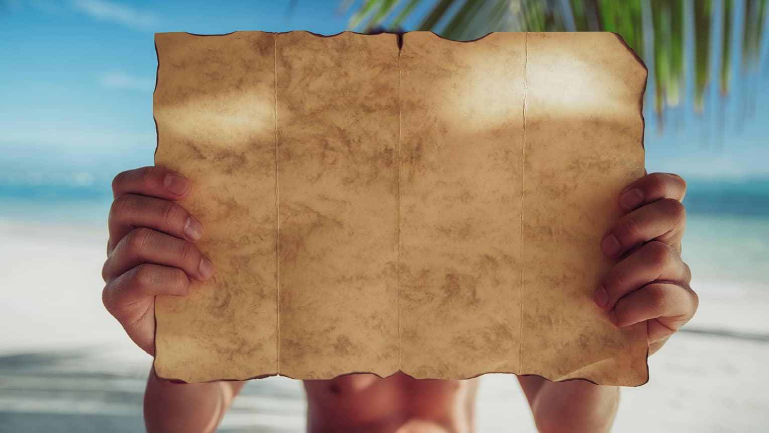 A close-up of a piece of parchment paper, yellow with age and burnt around the edges, being examined on a sandy beach.