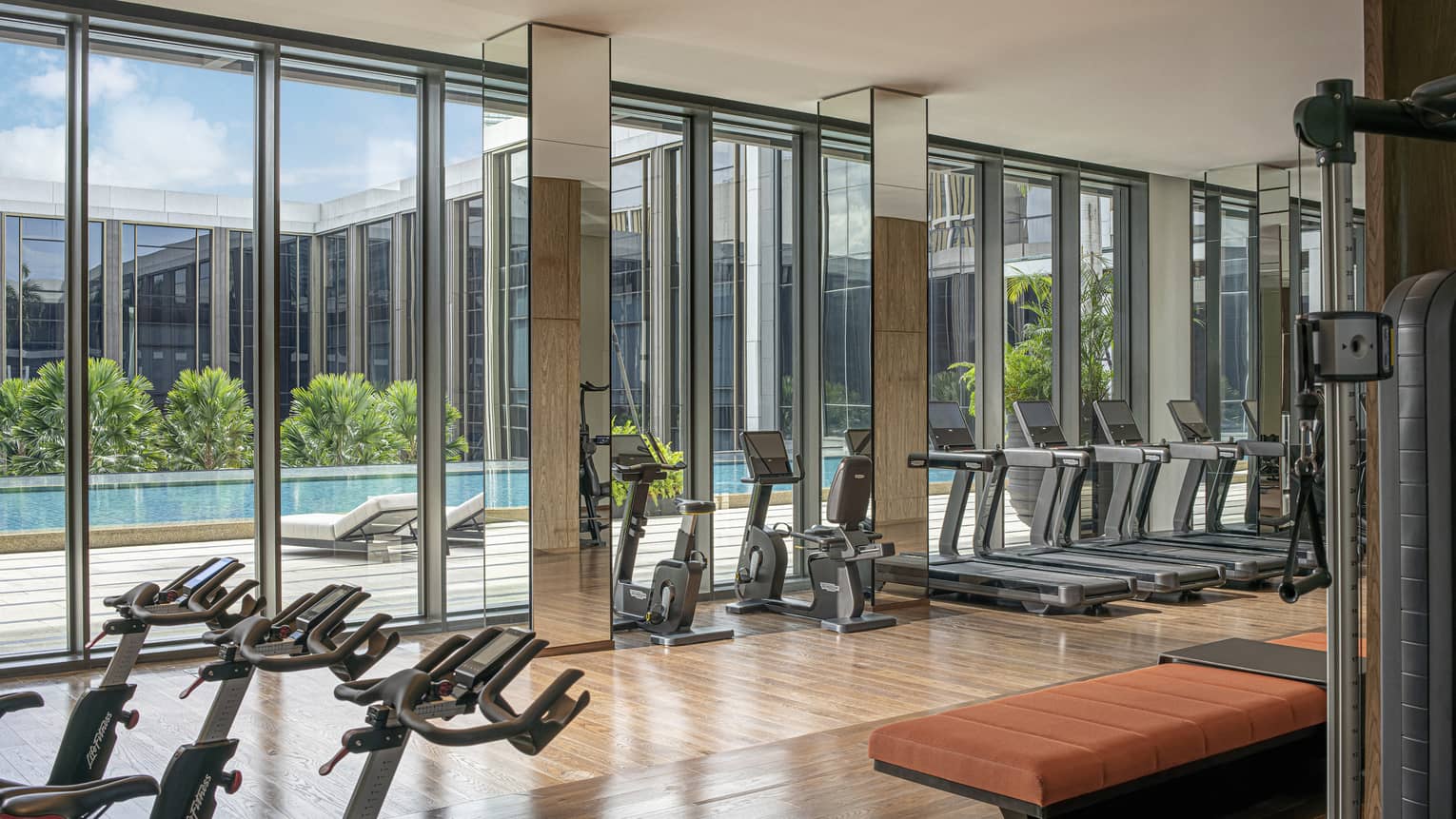 Hotel gym with floor-to-ceiling windows and rows of exercise equipment, natural light, pool views