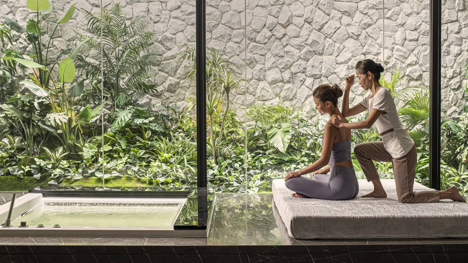 Person receiving a massage in a peaceful spa setting with large windows overlooking lush green plants and a small water feature.