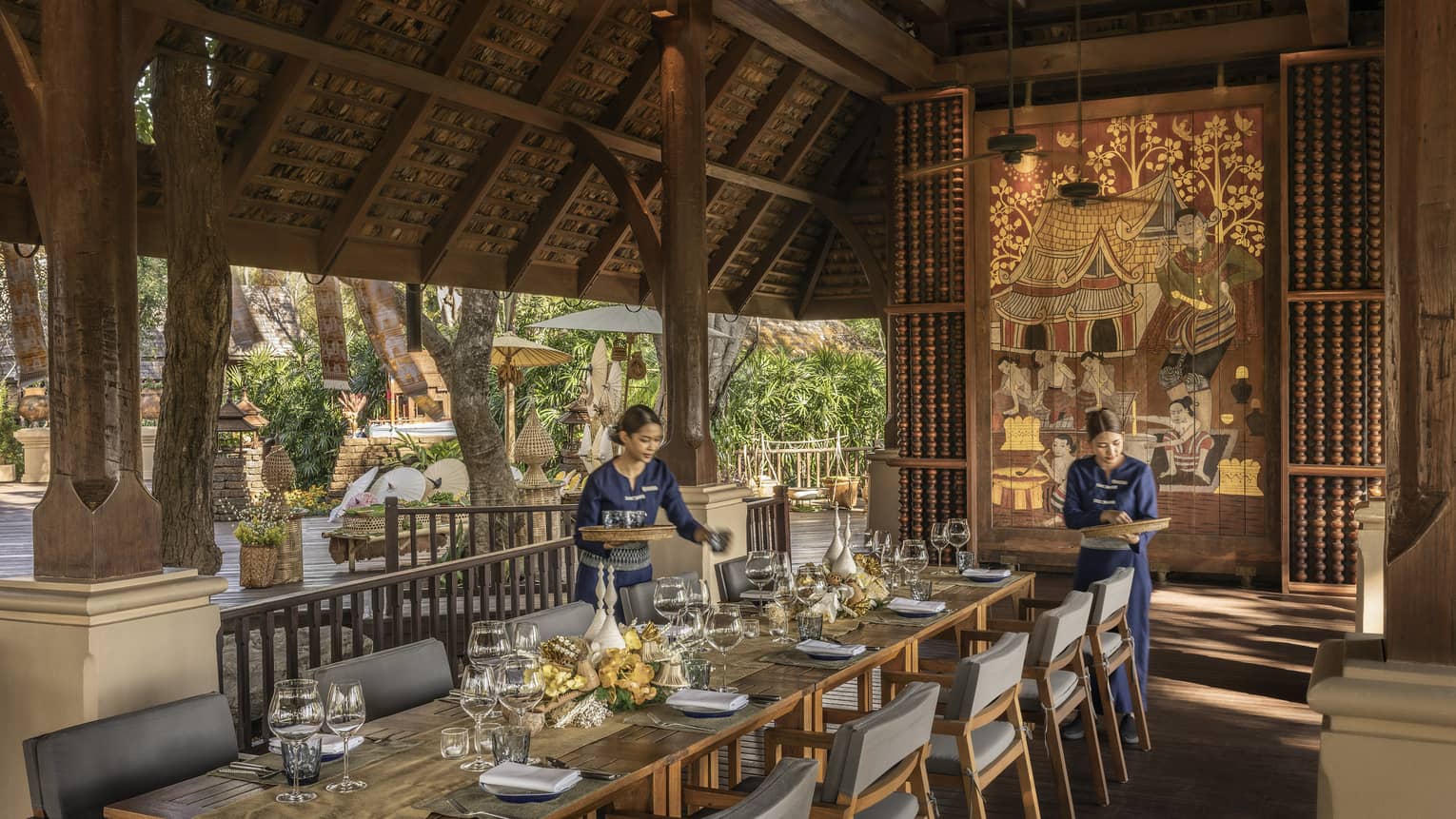 Two waitresses in blue set a long wooden table.