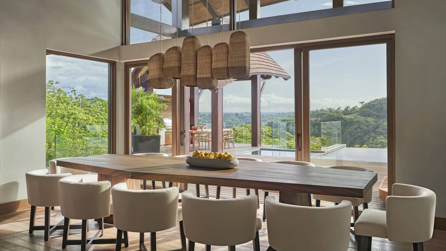 Dining room with long wooden table, 12 cream leather chairs, hanging wooden light fixture and wall of windows