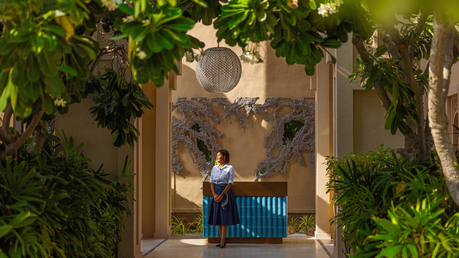Woman staff member wearing light-blue button down shirt and dark blue knee-length skirt stande next to a blue-tiled counter set surrounded by bright green plants and trees