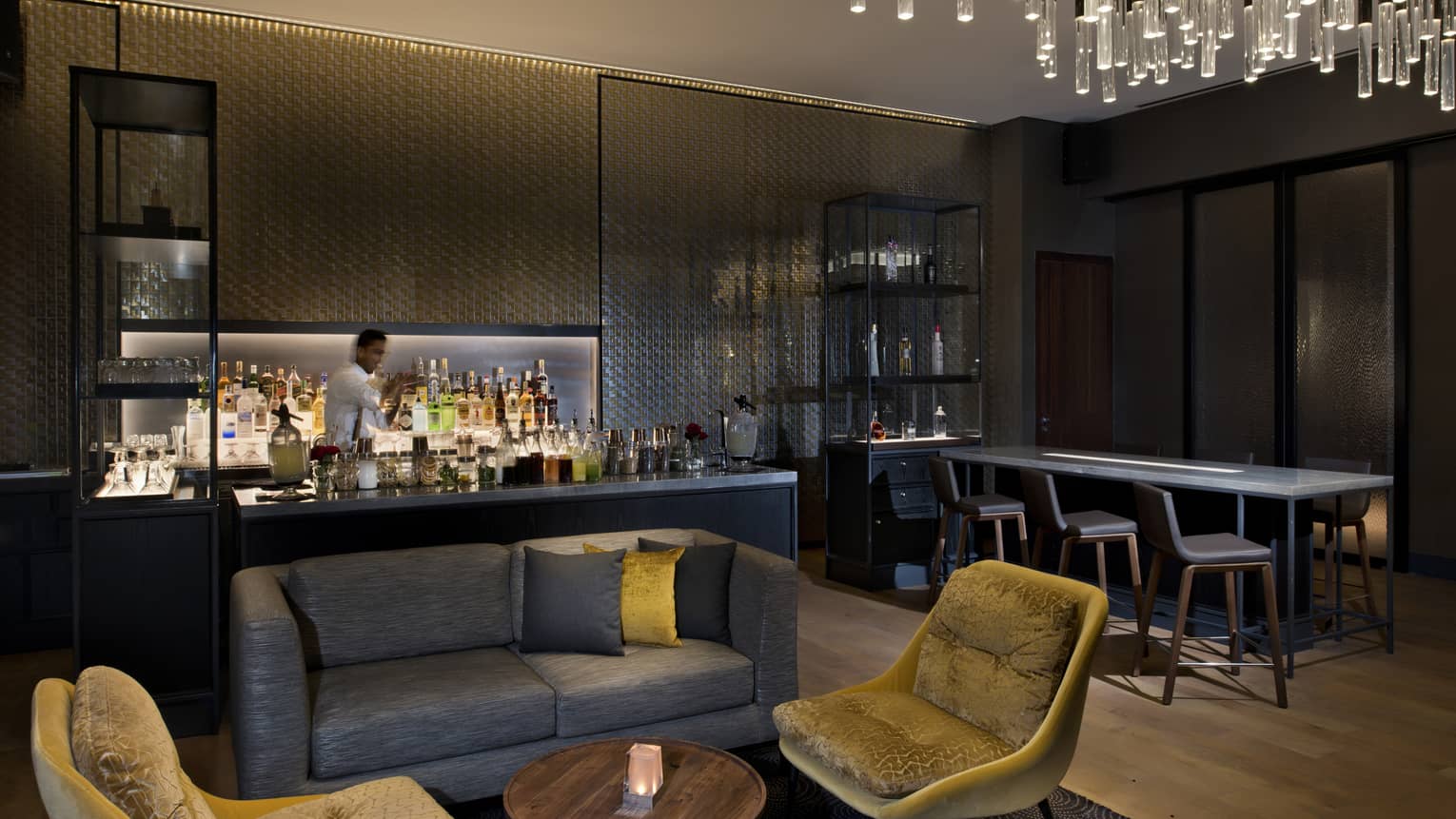 Dimly lit bar with bartender and lounge with brown loveseat and two velvet chairs under modern chandelier
