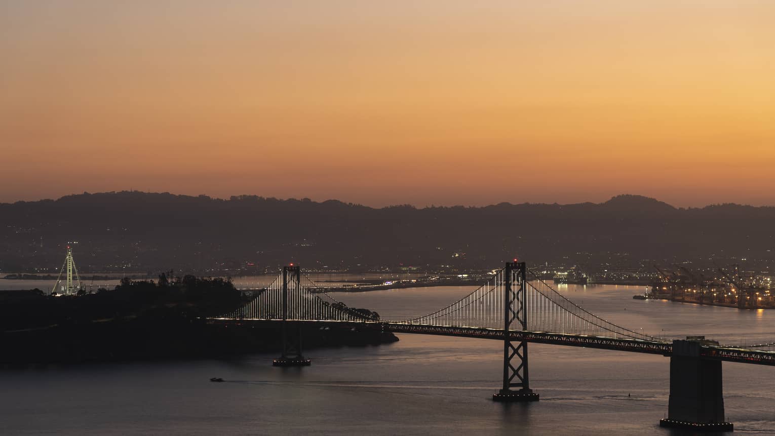A large bridge crossing a body of water with a city of lights and mountains behind it in the distance.