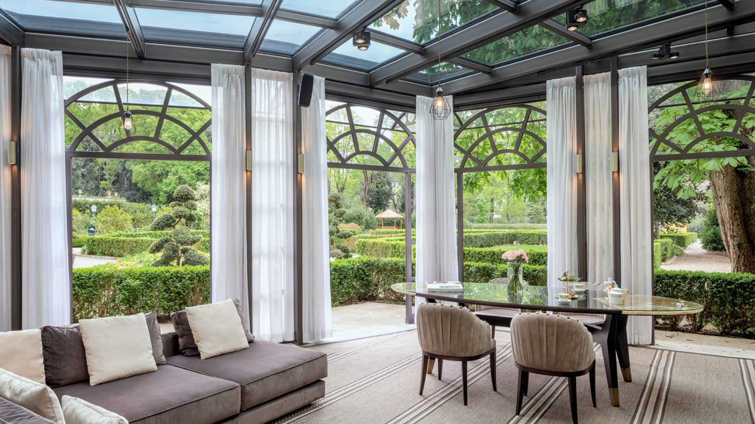Glass-enclosed living and dining room, surrounded by green gardens