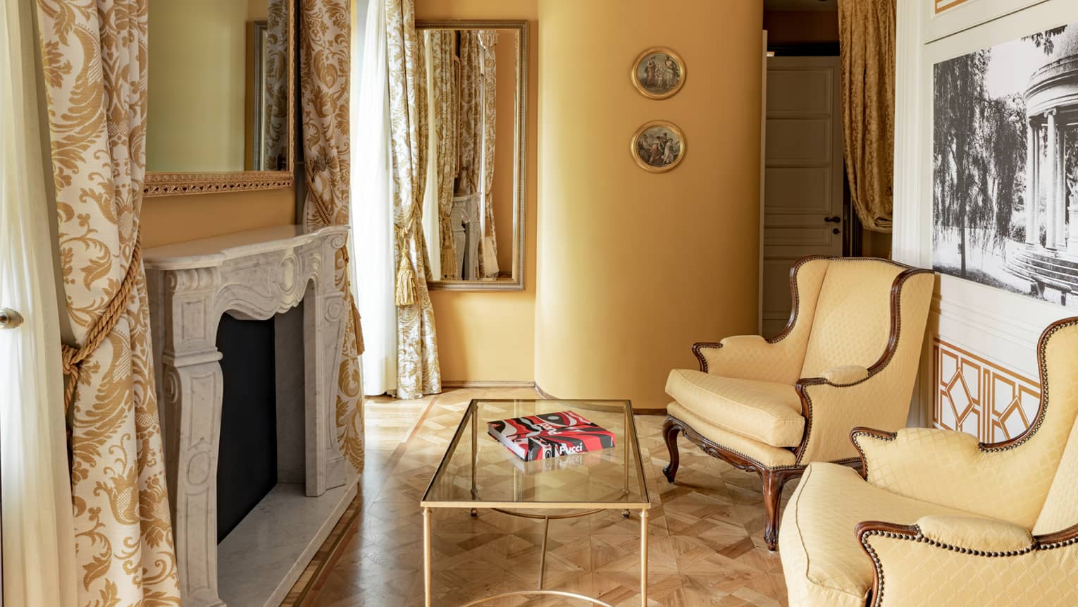 Sitting area with yellow walls, ornate fireplace, patterns drapery, two yellow wing-backed chairs