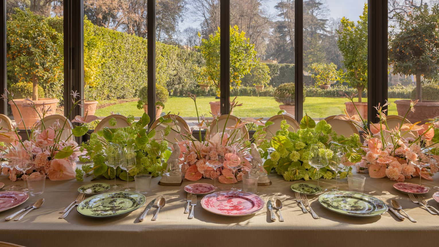 Bright banquet table set with pink and green plate and flowers beside tall windows looking out to manicured garden