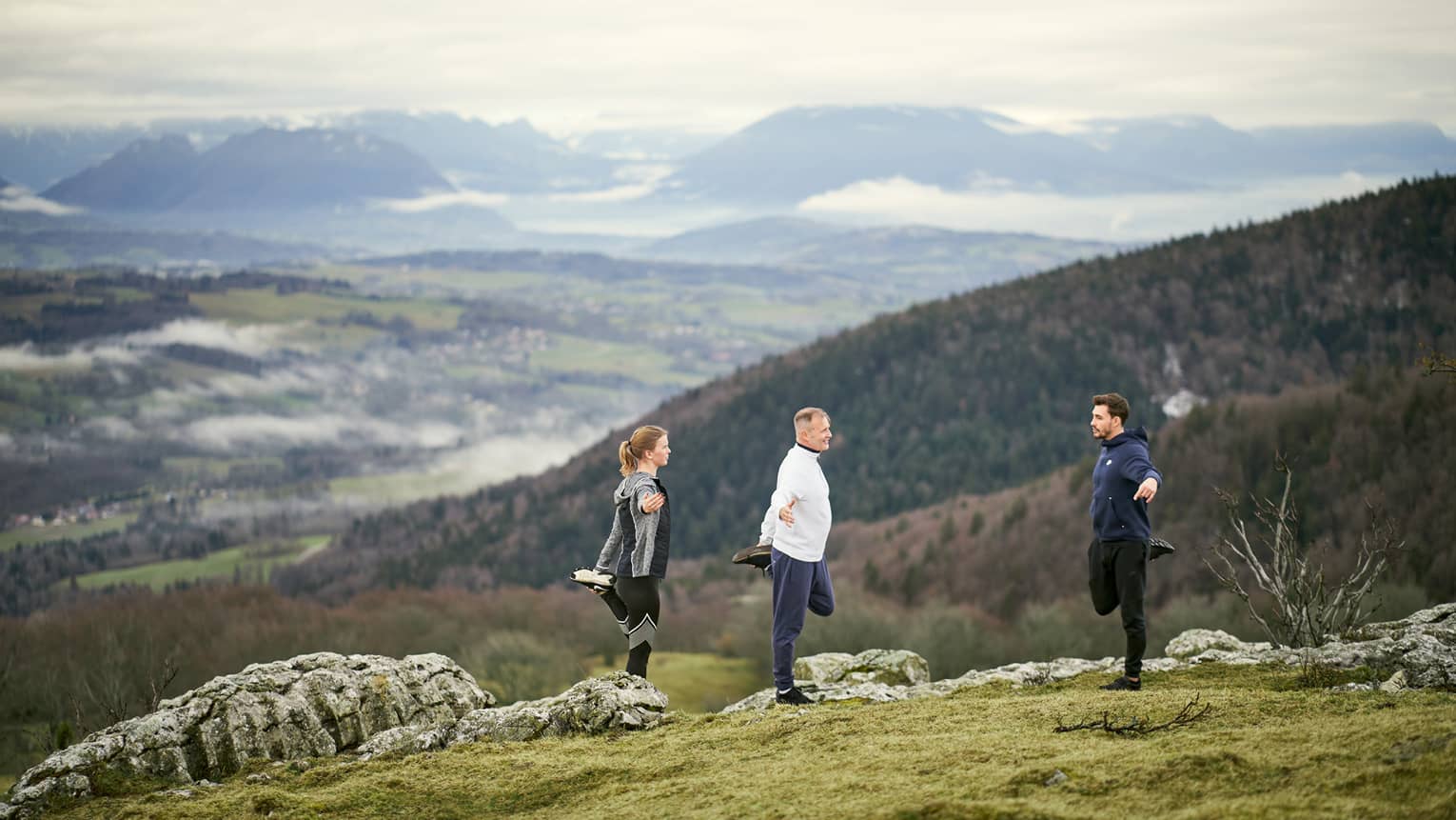 Woman and man standing on mountainside with male trainer, all stretching left quads 
