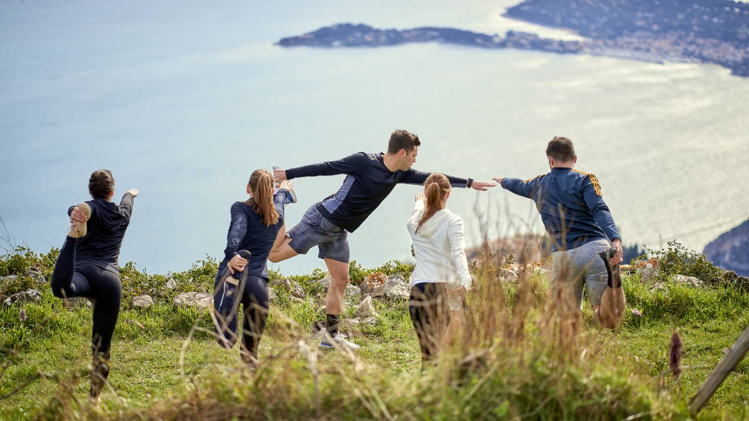 Rear view of four people in yoga poses on grassy cliff with instructor in front, sea view