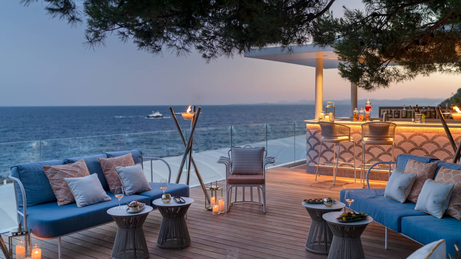 Club Dauphin wooden terrace, blue love seats, orange pillows, candles, torch, uplit bar, sea view at sunset