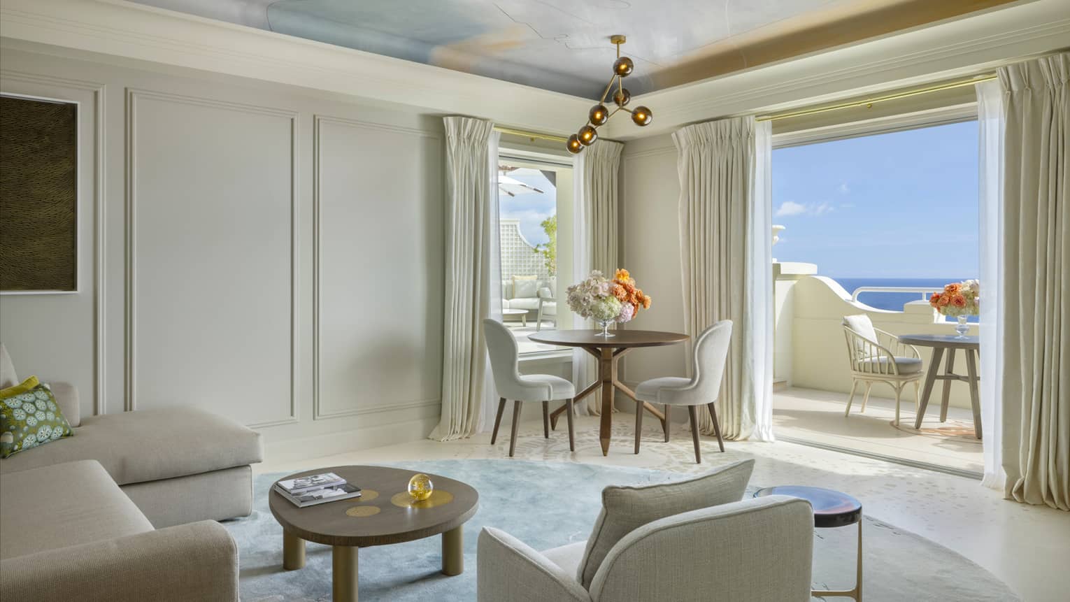Elegant hotel suite living room with cream-coloured sofa and arm chair, small dining table with two chairs, and sea-view terrace
