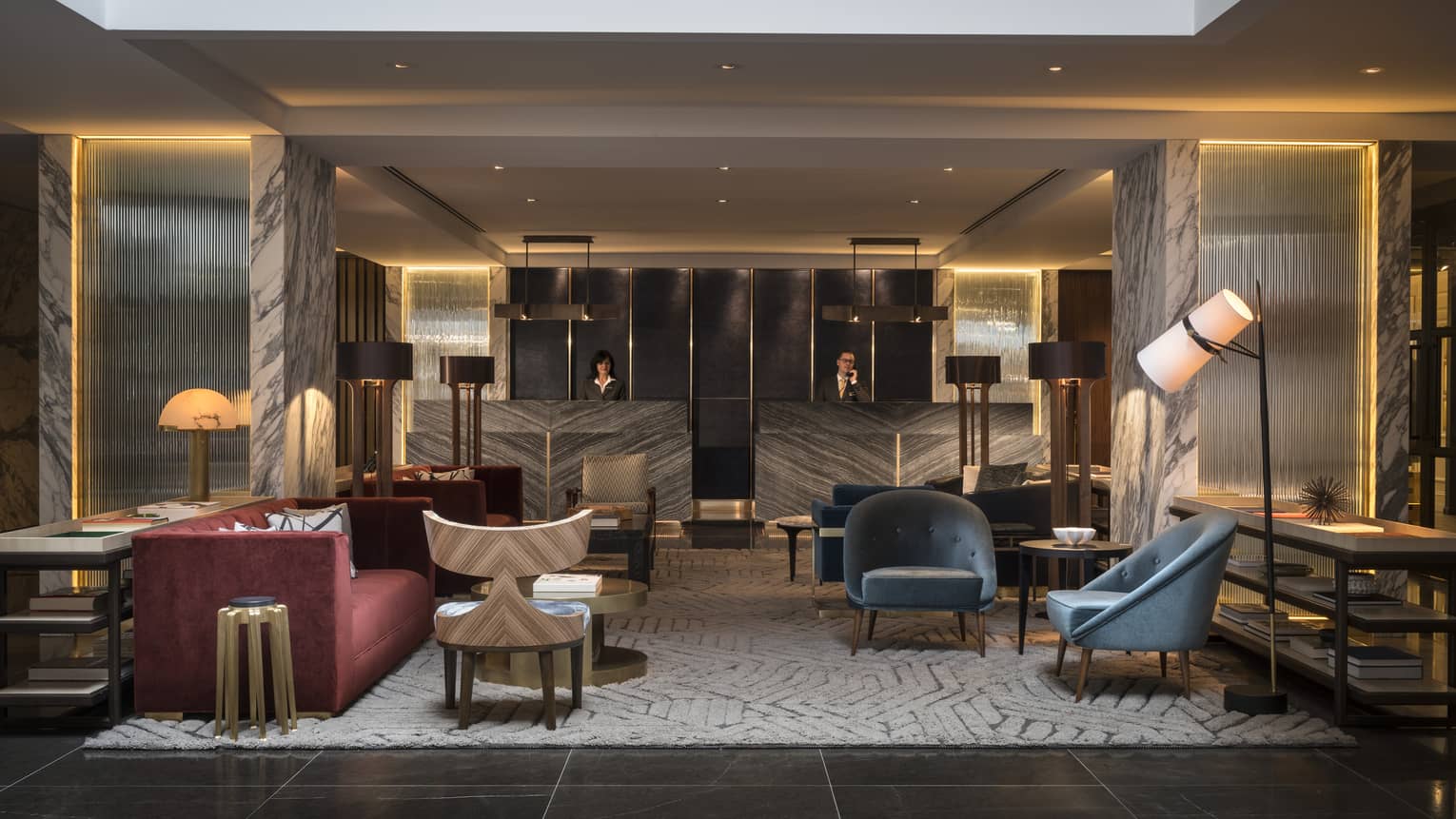 The Lobby of Four Seasons Houston is furnished with two blue velvet arm chairs, a gray and cream abstract rug, a red velvet couch, and an industrial black iron lamp