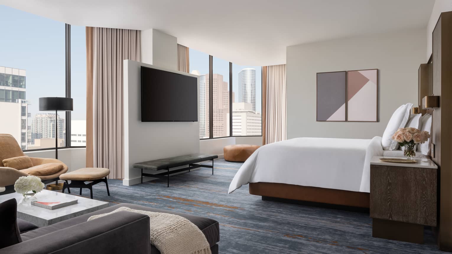 Spacious hotel bedroom with wall of windows, hanging TV, blue carpet, lounge seating and white king bed