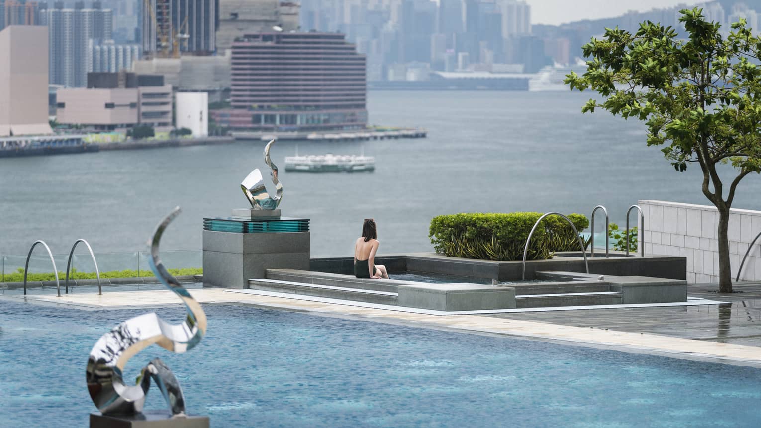 Woman in swimsuit sits on edge of swimming pool on patio near modern silver sculptures, waterfront