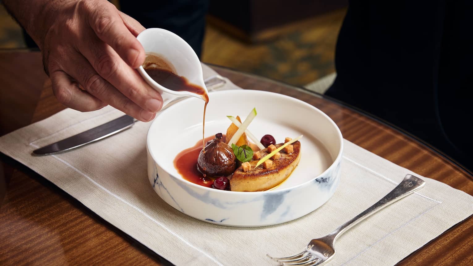 Chef pouring sauce over a gourmet dish with foie gras, fig and apple garnishes in a marble-patterned bowl.