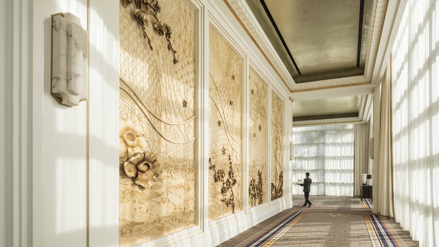 Hotel staff carries tray at end of long hallway past decorative paneled walls