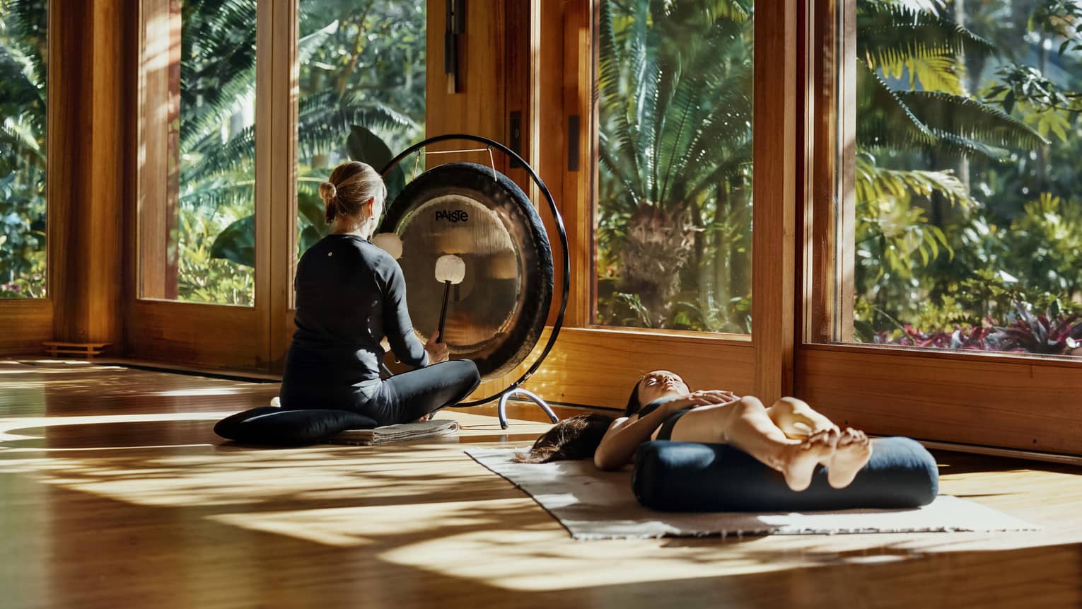 During a sound bath, a meditator lays on a mat, legs resting on a bolster, as a practitioner plays a gong with two mallets. 