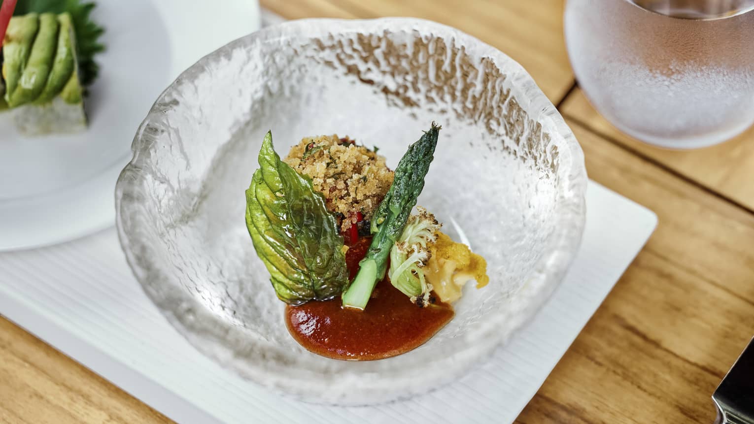 A croquette rests in a textured crystal bowl with red sauce, small grilled vegetables and a translucent fried herb. 