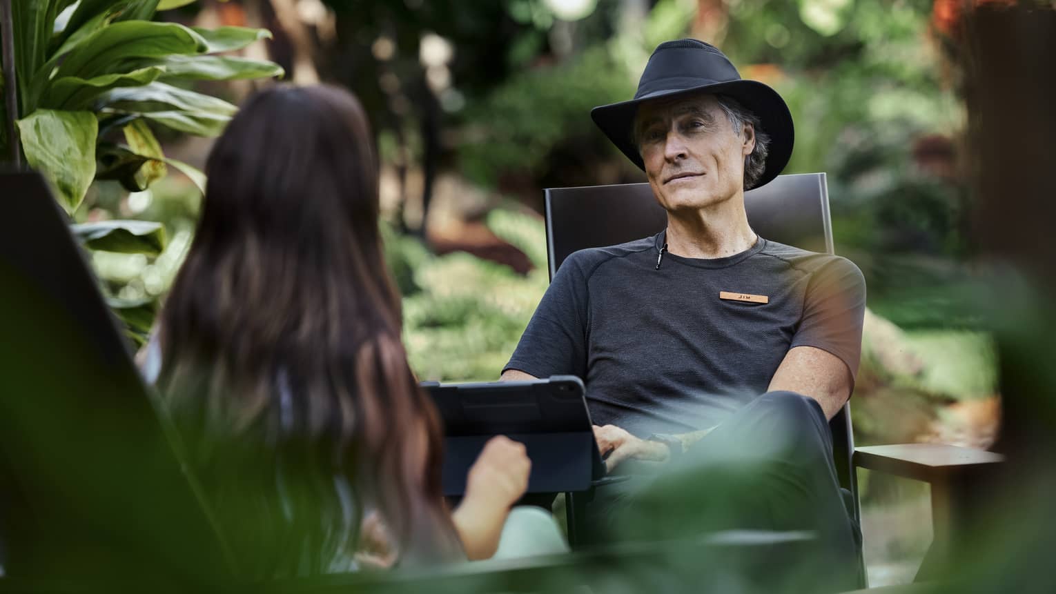Sitting amid lush vegetation, two people engage in conversation, one with kind eyes and a soft smile typing on a tablet.