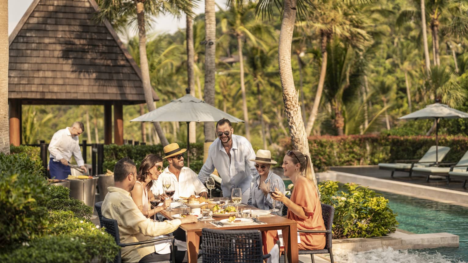 Group of laughing friends enjoy a poolside barbecue lunch