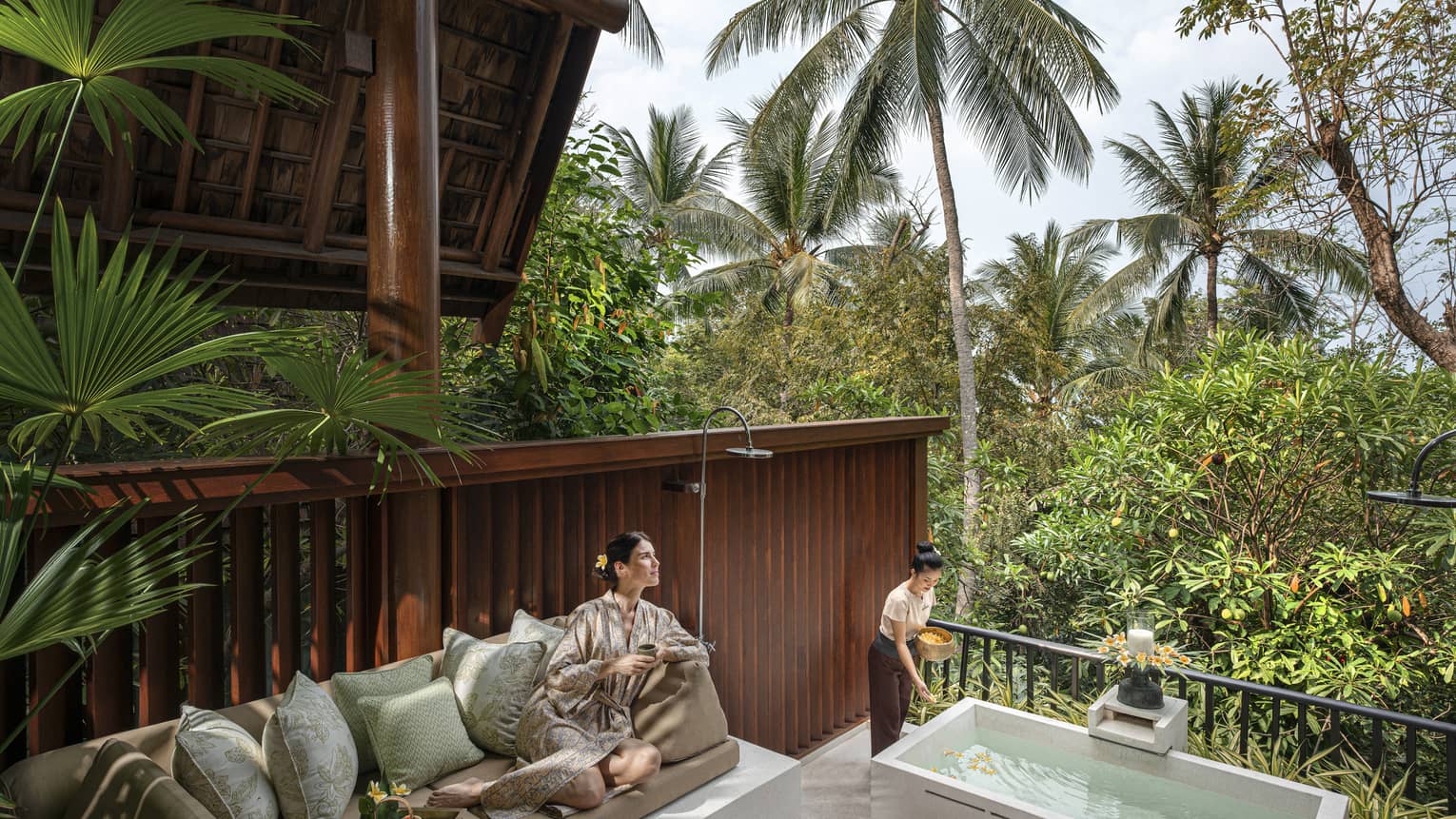 Woman in silk robe lounges on outdoor sofa as spa tech prepares bath