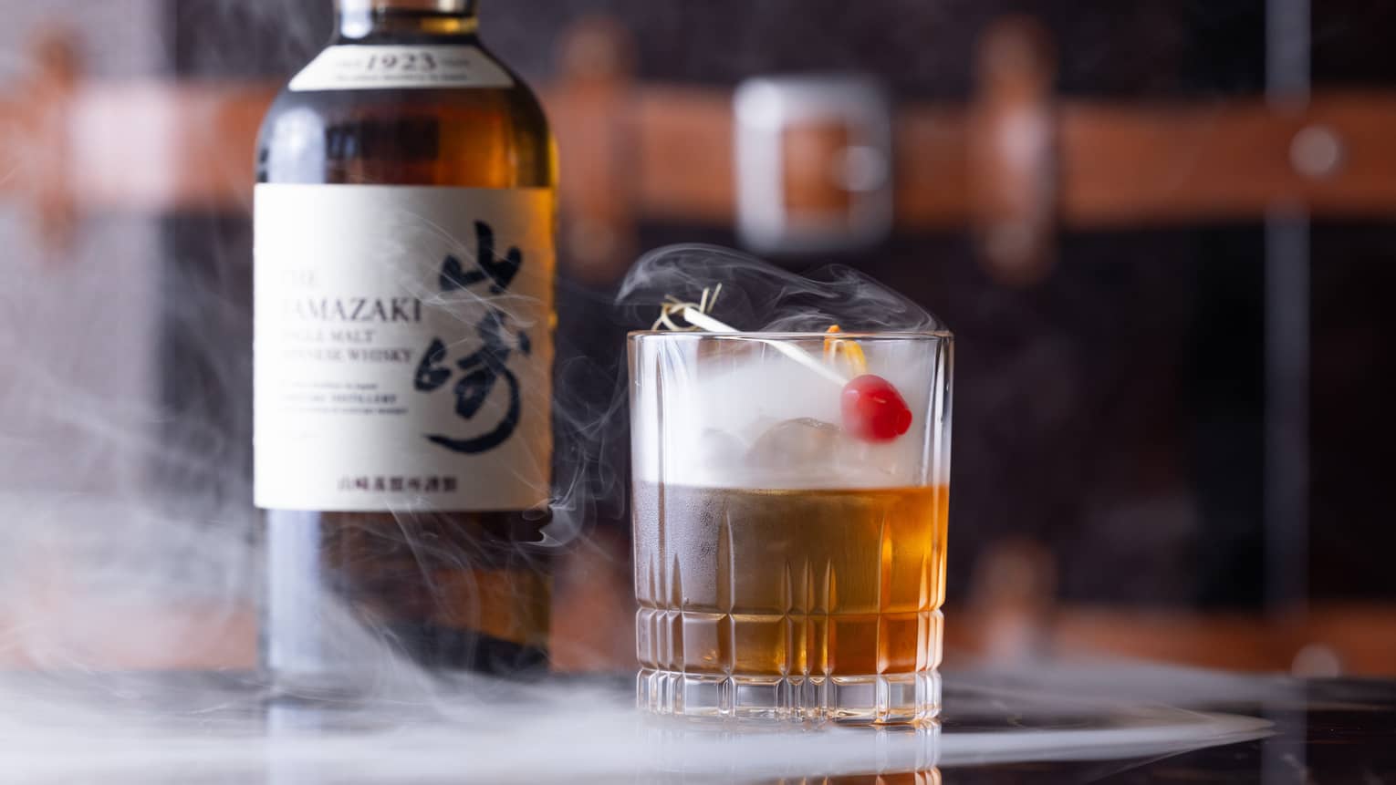 Bottle of Japanese whisky beside half-full cut-crystal glass with red cherry garnish, dry ice mist swirling from the glass.
