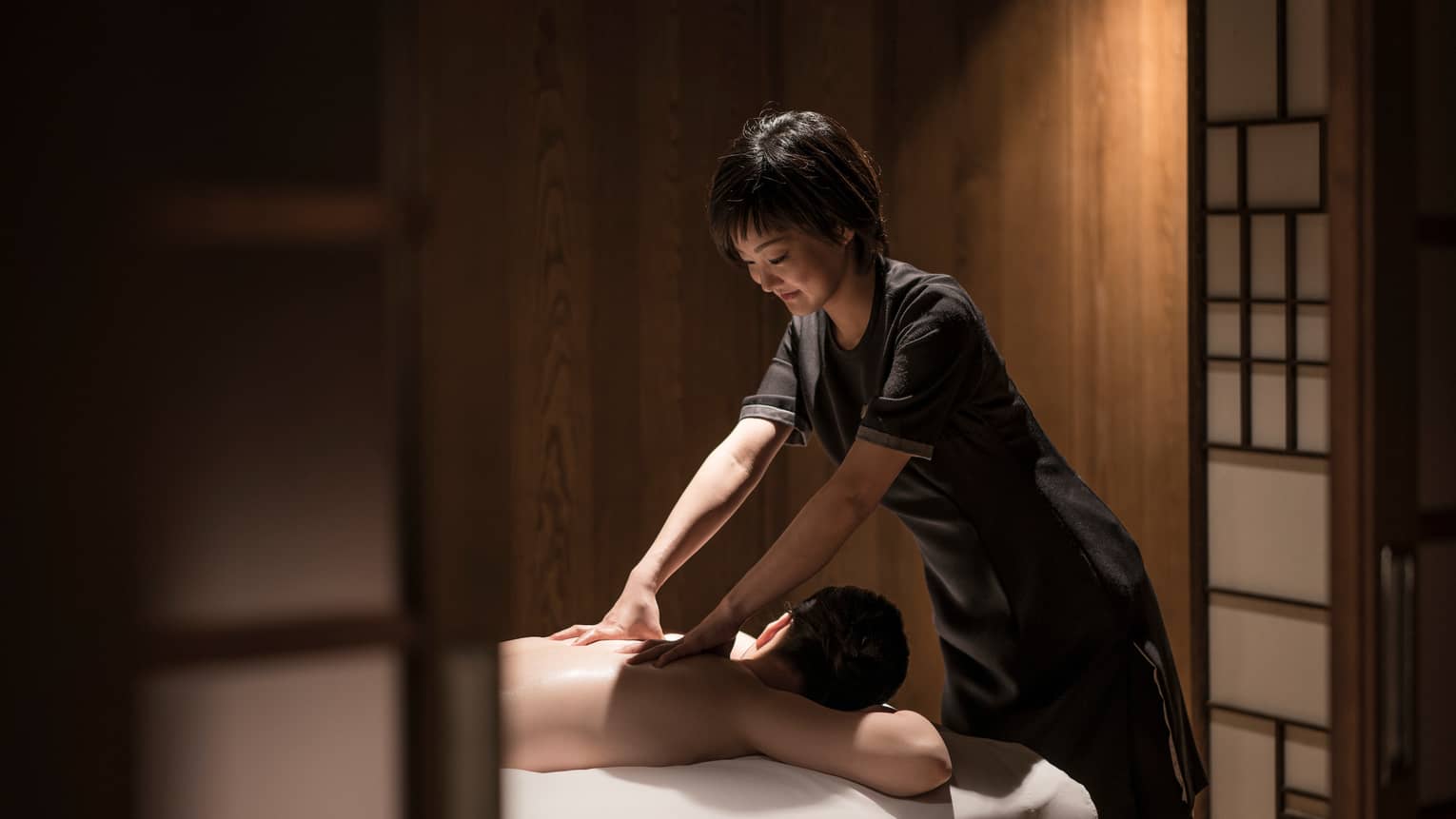 Man lies face-down on massage table as masseuse in black uniform rubs his shoulders