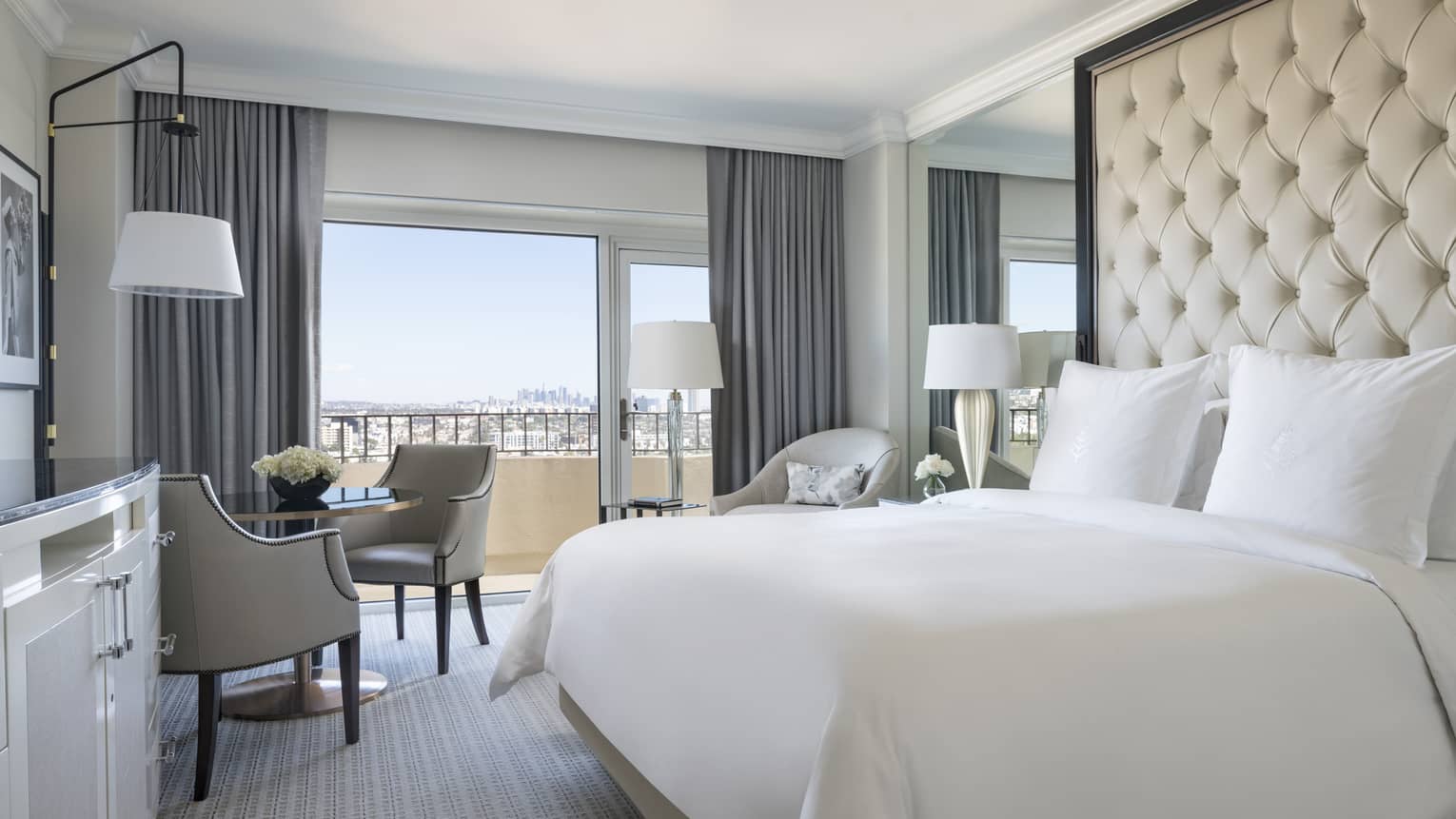 Premier Balcony Room hotel bed against padded, tufted wall, mirror, cafe table by sunny window
