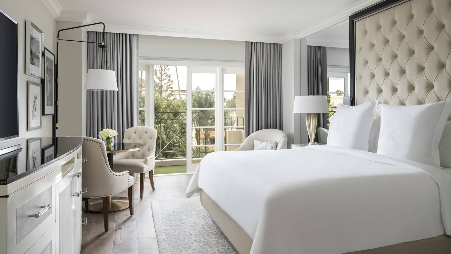A hotel room with a large bed with white sheets, black counter on white dresser, three chairs, and a large window.
