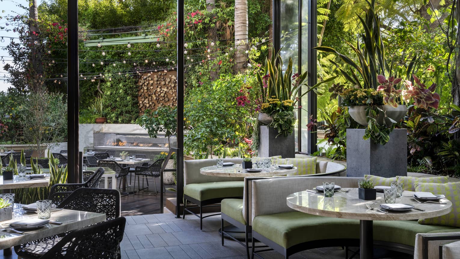 Restaurant seating with green chairs and many plants, with large windows leading to outdoor seating.