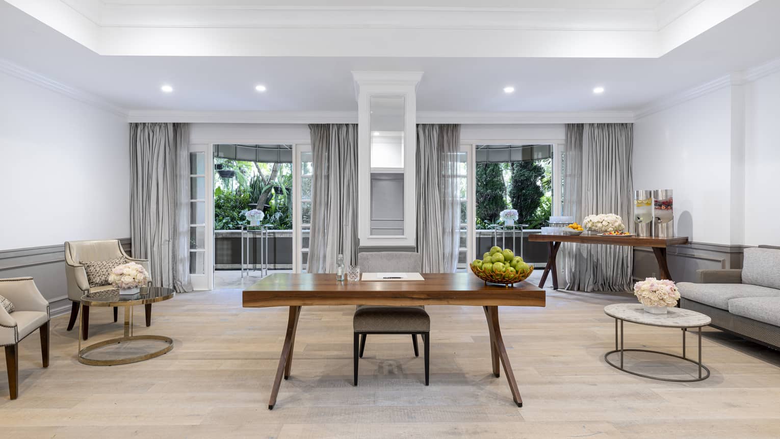 A seating room with a desk, two chairs, a sofa, a table with refreshments and large windows.