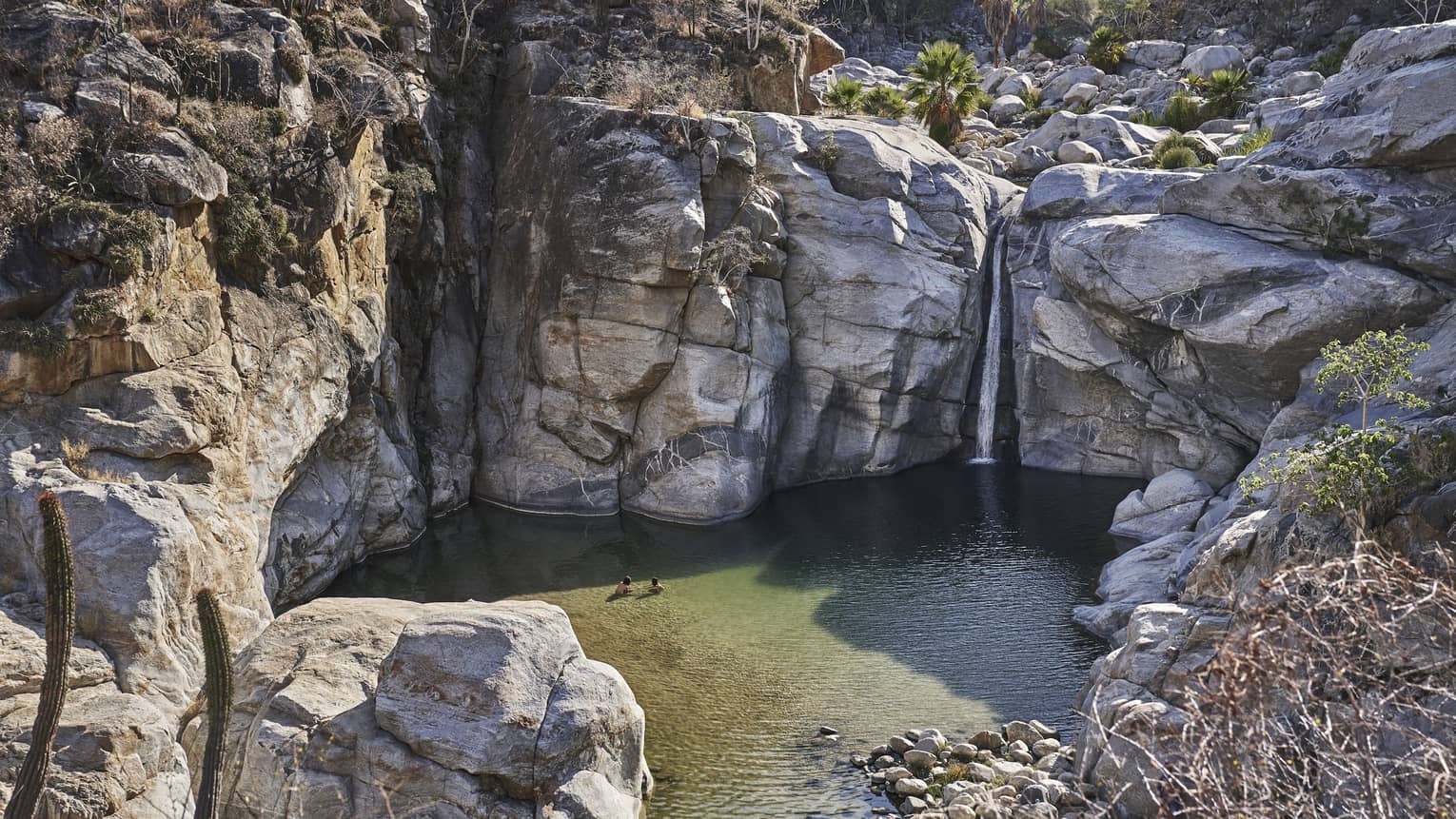 Two people wade in a small ocean lagoon surrounded by large cliffs, creating a serene and picturesque scene with clear water and rugged natural beauty.