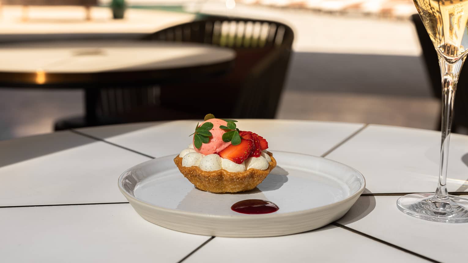 A white plate on a white table holds a dessert consisting of a base or shell, a white filling and a red fruit topping and is set next to a pale drink in a clear stemmed glass