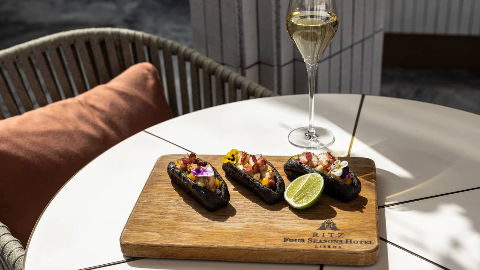 Three colorful food items on a wood serving board next to a pale drink in a clear stemmed glass, all on a white table