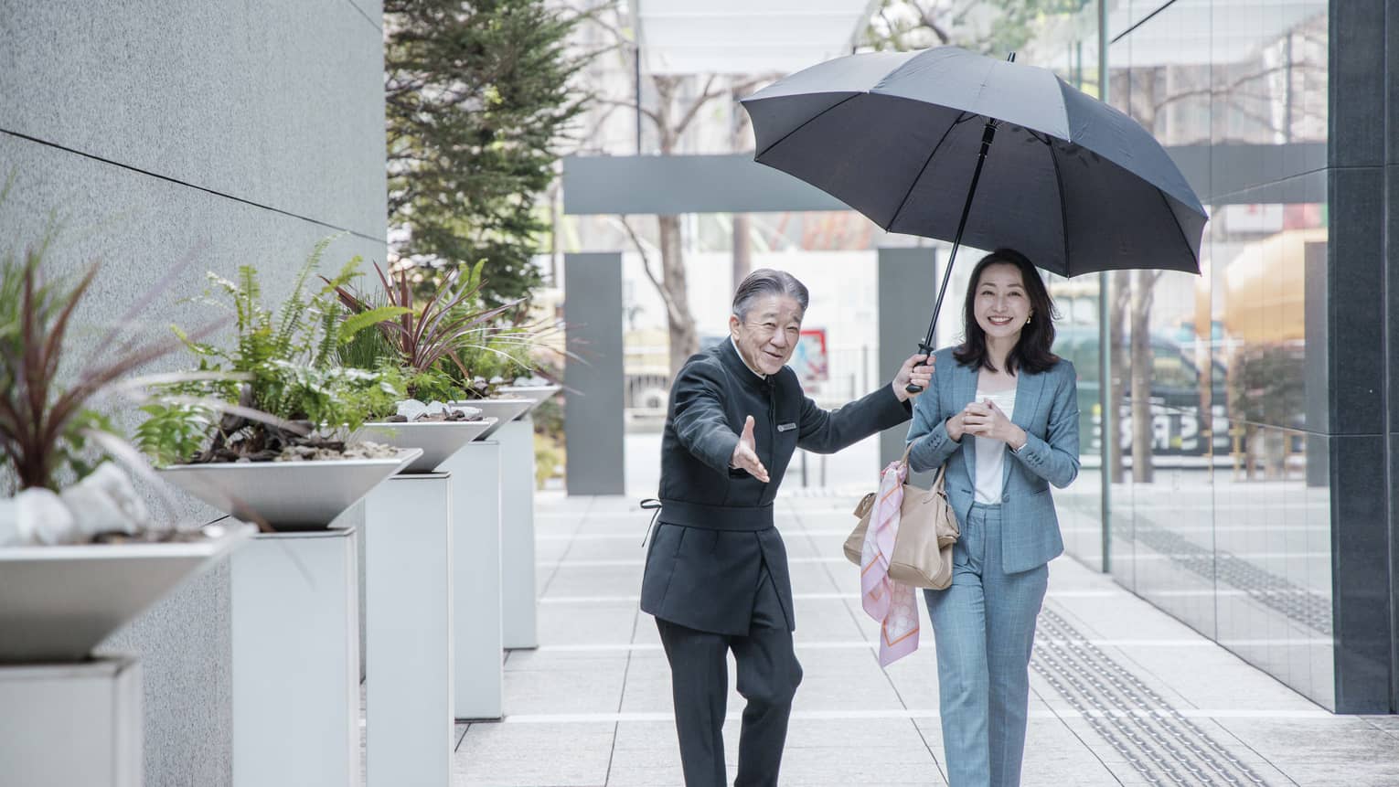 Staff member leads smiling guest down walkway while sheltering them with large black umbrella